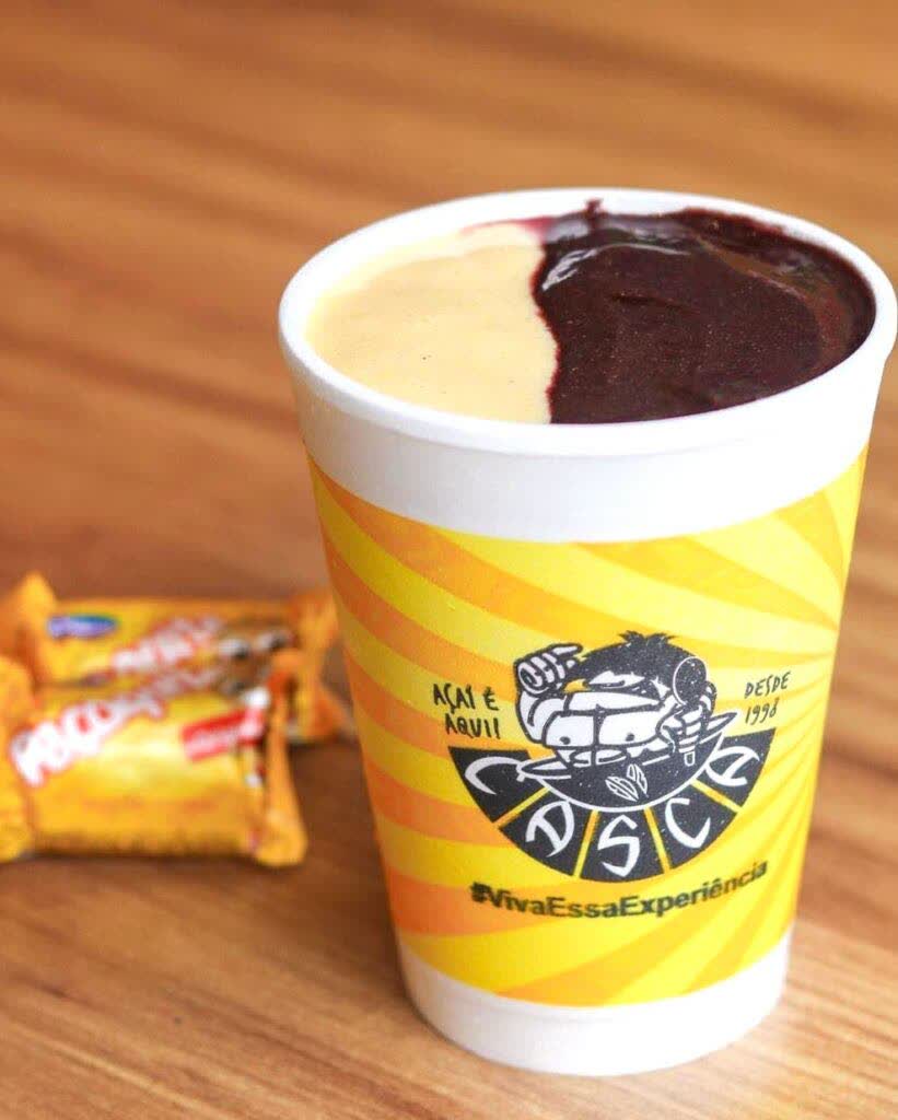 Copo oficial do Casca Açaí com açaí e creme claro, ao lado de pacotes de paçoca.